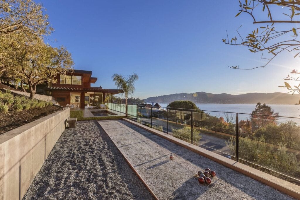 A Modern House With Large Windows Overlooking A Lake And Mountains. In The Foreground, There'S A Bocce Ball Court Surrounded By A Glass Fence. The Sun Casts Long Shadows, Highlighting The Lush Greenery And Clear Blue Sky.