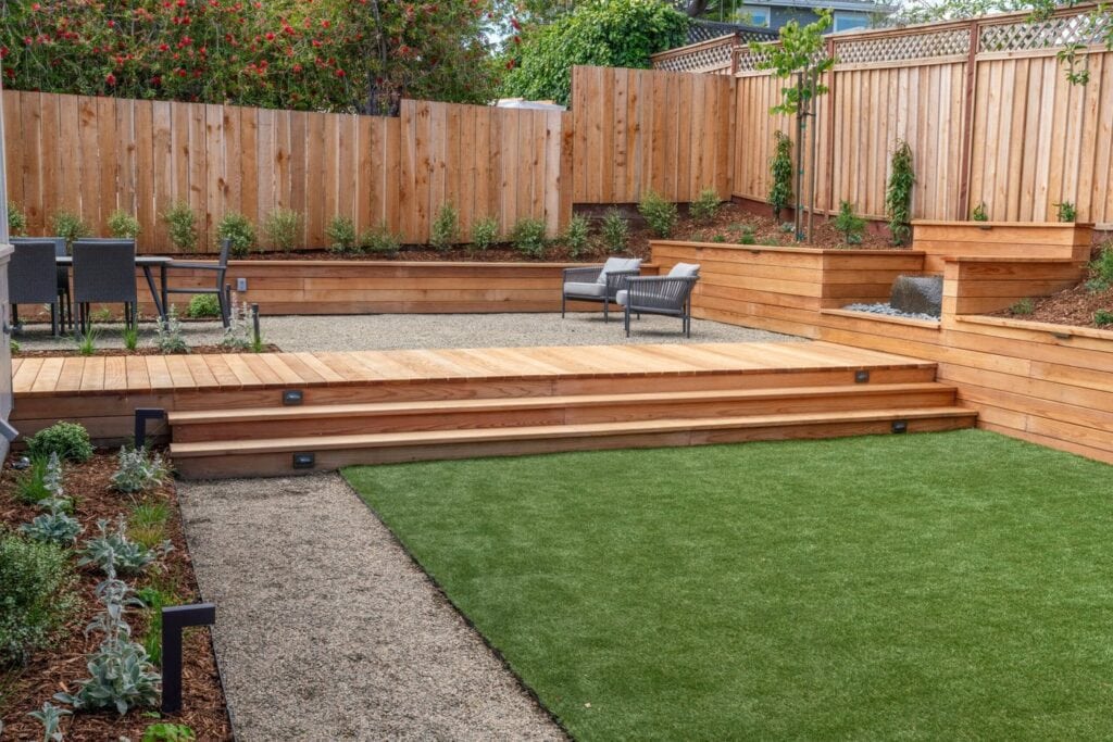 A Landscaped Backyard With A Wooden Deck, Steps, And Seating Area. The Yard Features A Lawn, Gravel Pathways, And Raised Garden Beds Enclosed By A Wooden Fence. Some Greenery Is Planted Along The Perimeter And On The Slope.