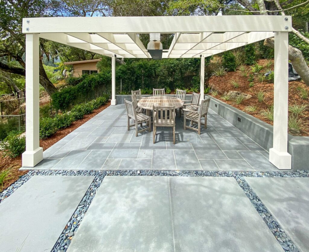 A Patio With A Wooden Pergola Features An Outdoor Dining Set On Stone Tiles. The Setting Is Surrounded By Greenery And A Landscaped Garden.