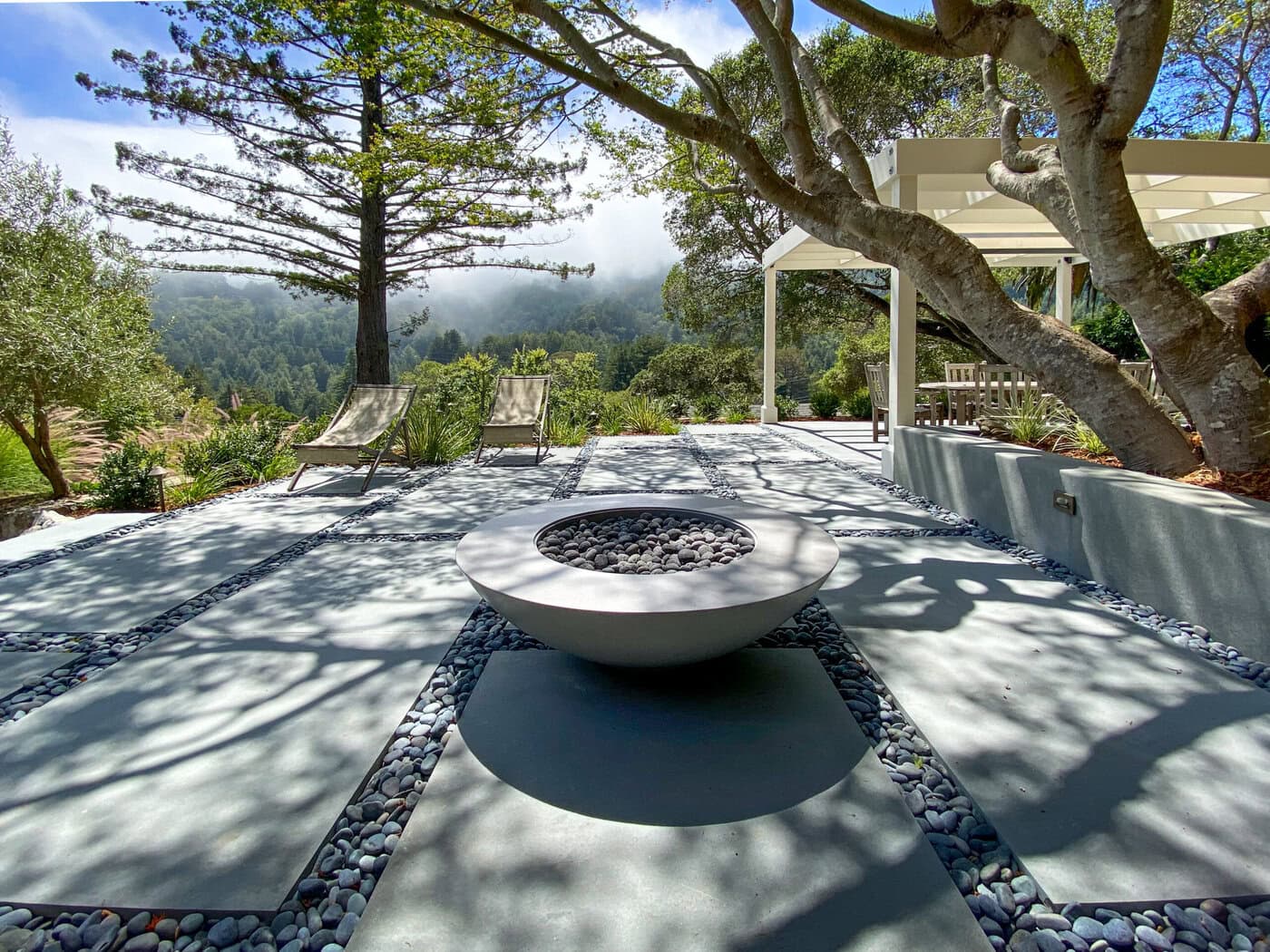 A Serene Patio With Modern Lounge Chairs And A Circular Fire Pit On A Smooth Stone Surface. Surrounded By Lush Greenery And Trees, Overlooking A Misty Forested Landscape. Dappled Sunlight Creates Patterns On The Ground.