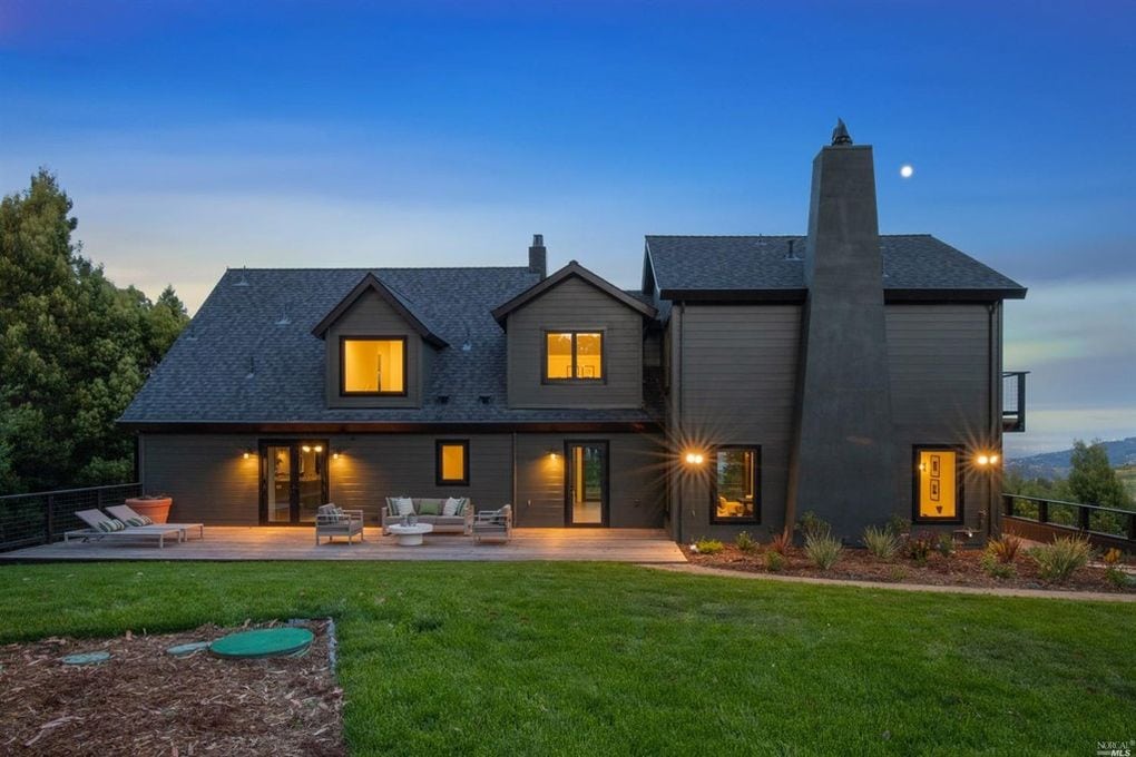 A Modern Two-Story House With Dark Exterior, Lit Warmly From Within. It Features Large Windows, A Spacious Wooden Deck With Patio Furniture, And Is Surrounded By A Well-Kept Lawn. The Evening Sky Is Clear, With A Small Moon Visible.