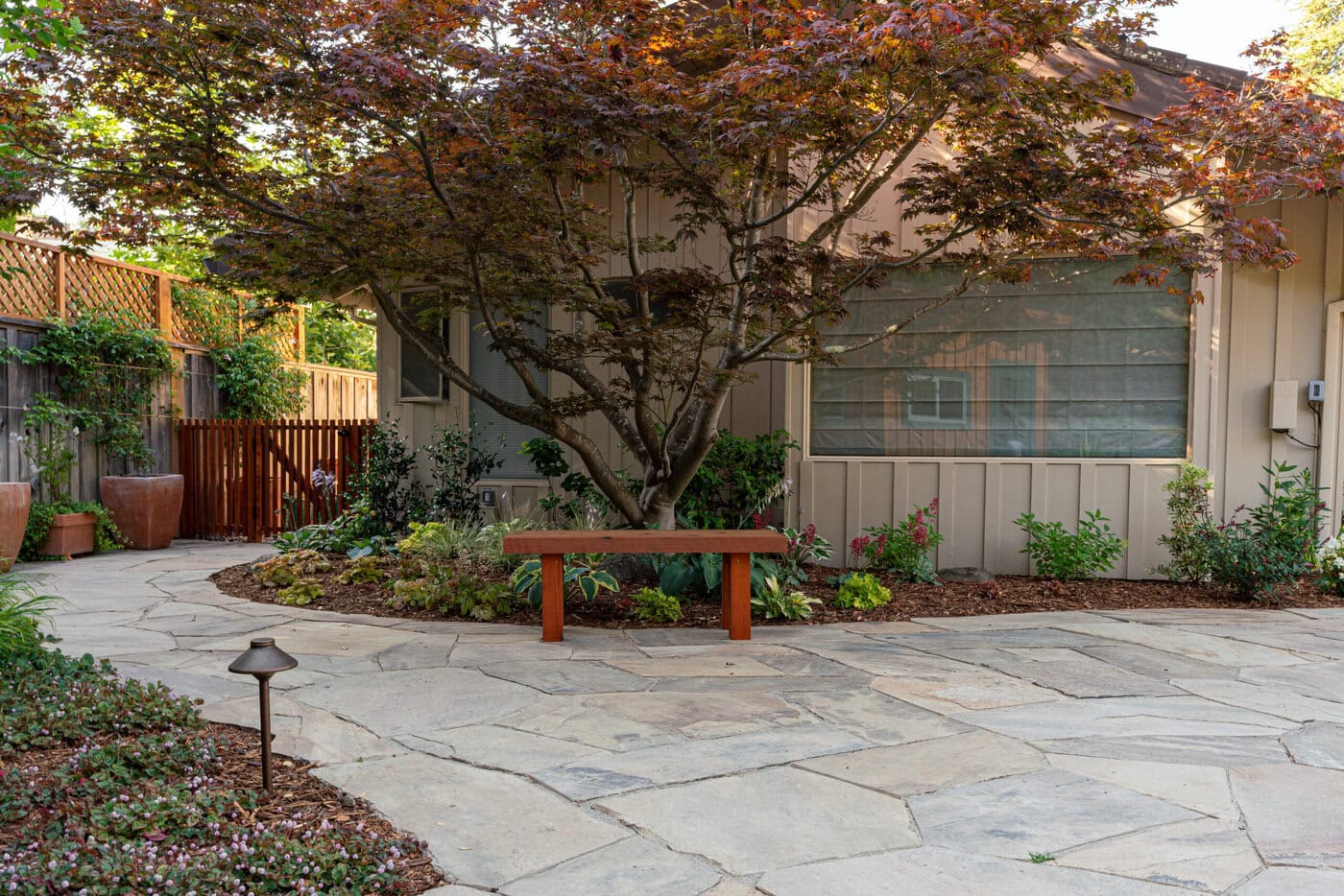 A Tranquil Garden Scene With A Stone-Paved Path Leading To A Wooden Bench Shaded By A Large Tree. Lush Greenery And Potted Plants Surround The Area. A Wooden Fence And A House With Large Windows Are In The Background.