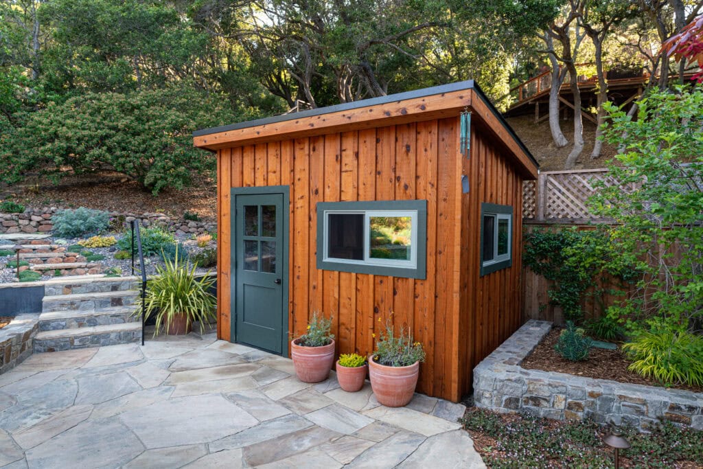 A Small, Modern Wooden Shed With A Slanted Roof Sits In A Landscaped Backyard. It Features A Green Door And Rectangular Windows. Three Large Plant Pots Are Placed In Front, And Stone Steps And Foliage Are Visible In The Background.