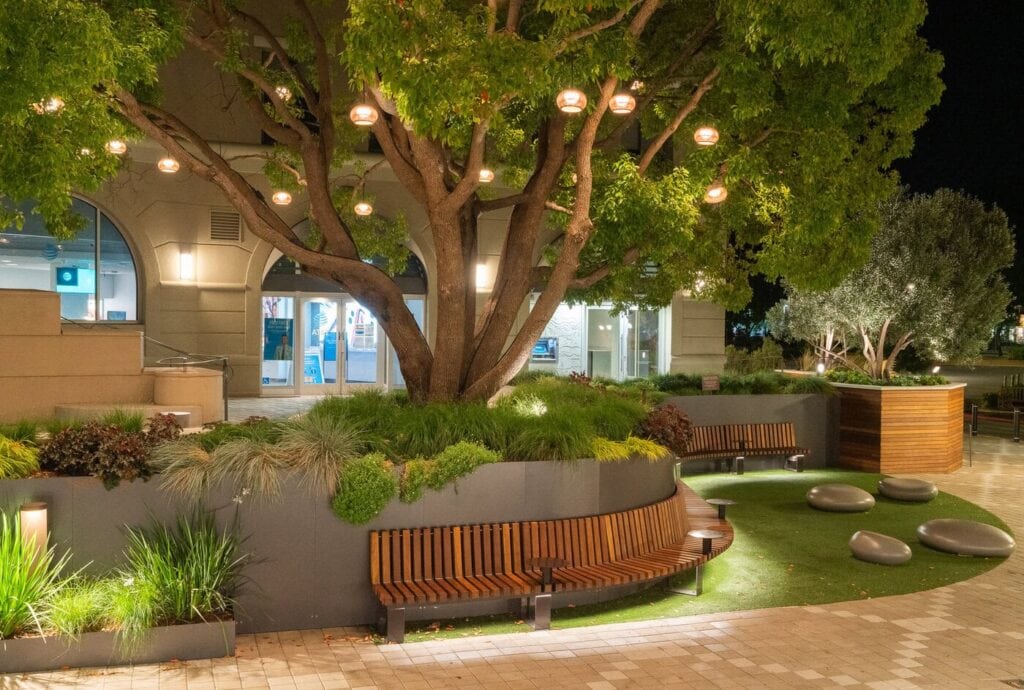 A Night Scene Of A Tranquil Outdoor Courtyard Featuring A Large Tree Adorned With Glowing Lights. Surrounding It Are Wooden Benches, Green Shrubs, And A Manicured Lawn With Stone Sculptures, Creating A Serene Ambiance.