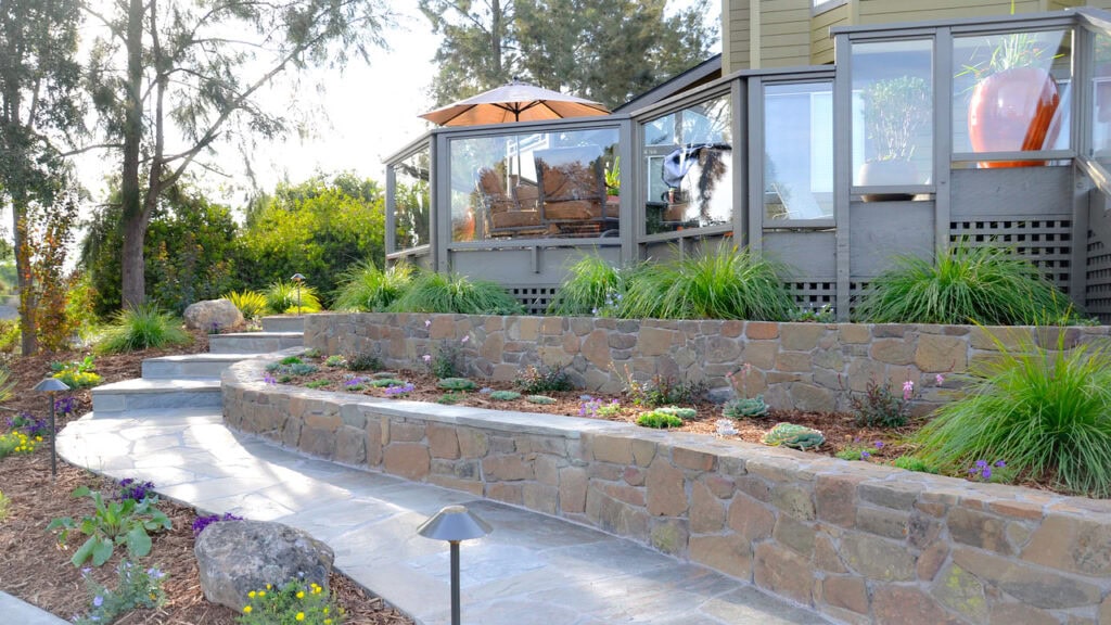 A Terraced Garden With Stone Retaining Walls And Lush Greenery Leads To A Modern Glass-Enclosed Patio. A Curved Stone Path Is Lined With Plants And A Small Outdoor Light, With Trees In The Background.