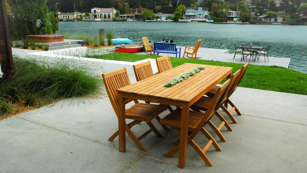 Wooden Dining Table With Chairs Set On A Patio Overlooking A Lake. The Scene Includes Lush Green Grass, A Dock With Seating, And Various Boats Near The Water. Houses And Trees Line The Opposite Shore Under A Partially Cloudy Sky.