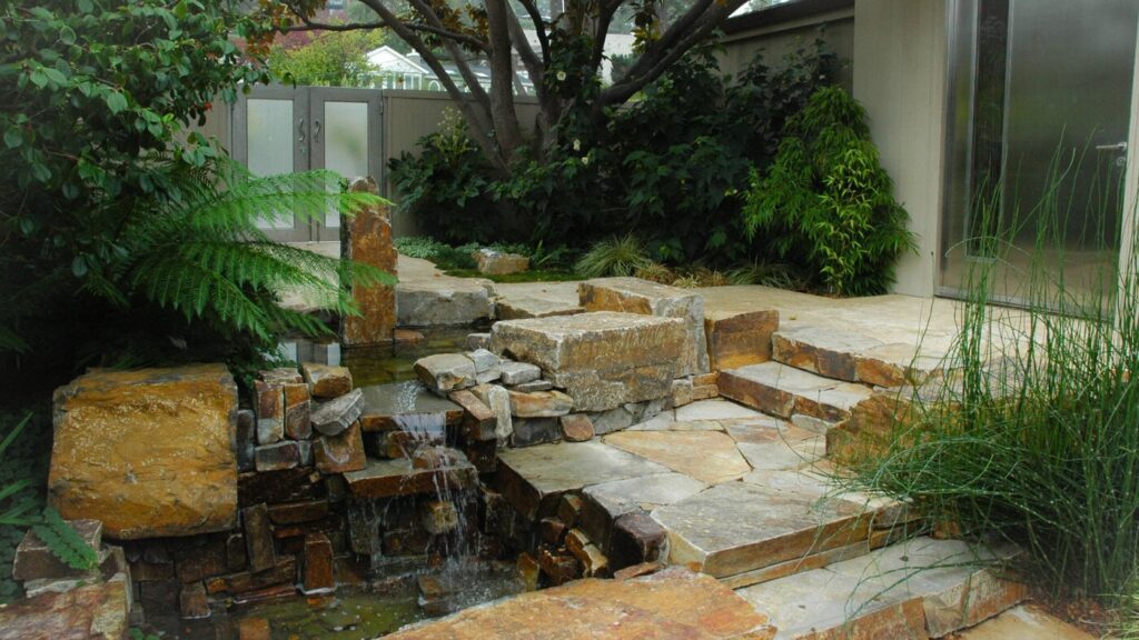 A Tranquil Garden Scene With A Small Cascading Waterfall Flowing Over Stacked Stone Slabs Into A Pond. Lush Greenery, Including Ferns And Shrubs, Surrounds The Water Feature. Stone Steps Lead To A Door And A Partially Visible Building.