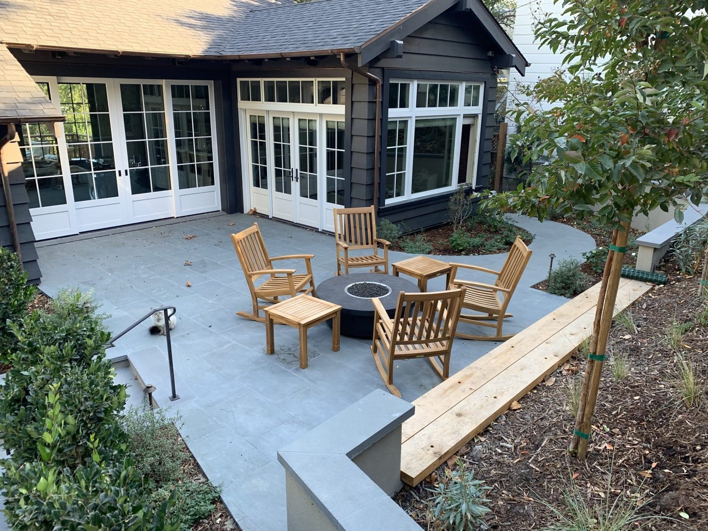 A Patio With Gray Stone Flooring Features A Circular Fire Pit Surrounded By Five Wooden Chairs And A Small Table. The Area Is Bordered By Greenery, And In The Background, There Are Large Windows And Doors Of A House.