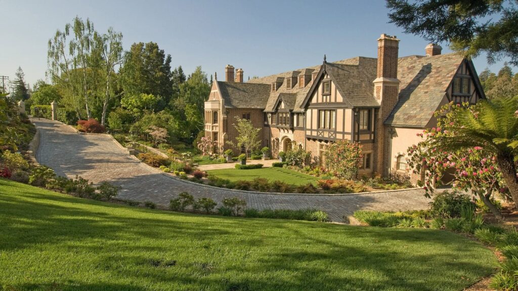 A Large, Elegant Tudor-Style Mansion With Brick And Timbering Is Surrounded By Lush Green Lawns And Gardens. A Cobblestone Driveway Curves In Front Of The House, And Tall Trees Frame The Scene Under A Clear Blue Sky.