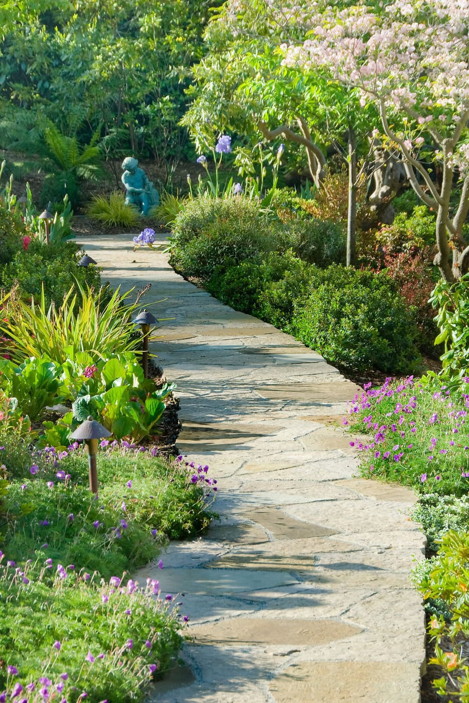 A Stone Path Winds Through A Lush Garden With Various Green Plants And Blooming Flowers. A Small Blue Statue Is Visible Among The Greenery, And A Tree With Pink Blossoms Provides Shade. The Scene Is Bright And Serene.