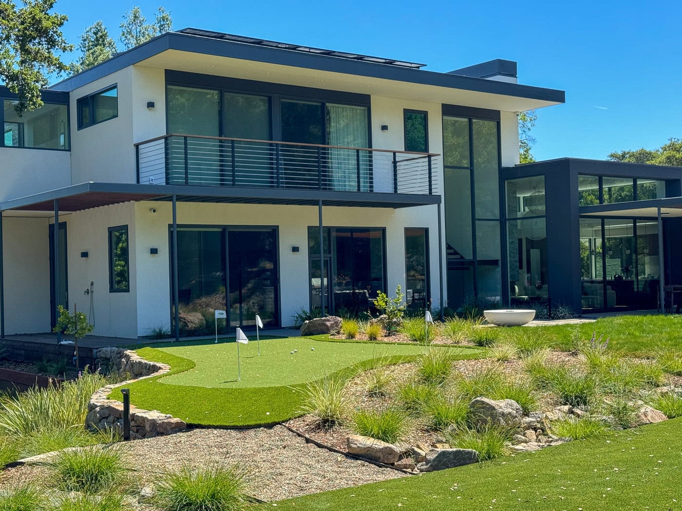 A modern two-story house with a spacious balcony and large glass windows. In the foreground is a small putting green surrounded by lush landscaping and rocky accents, under a clear blue sky.