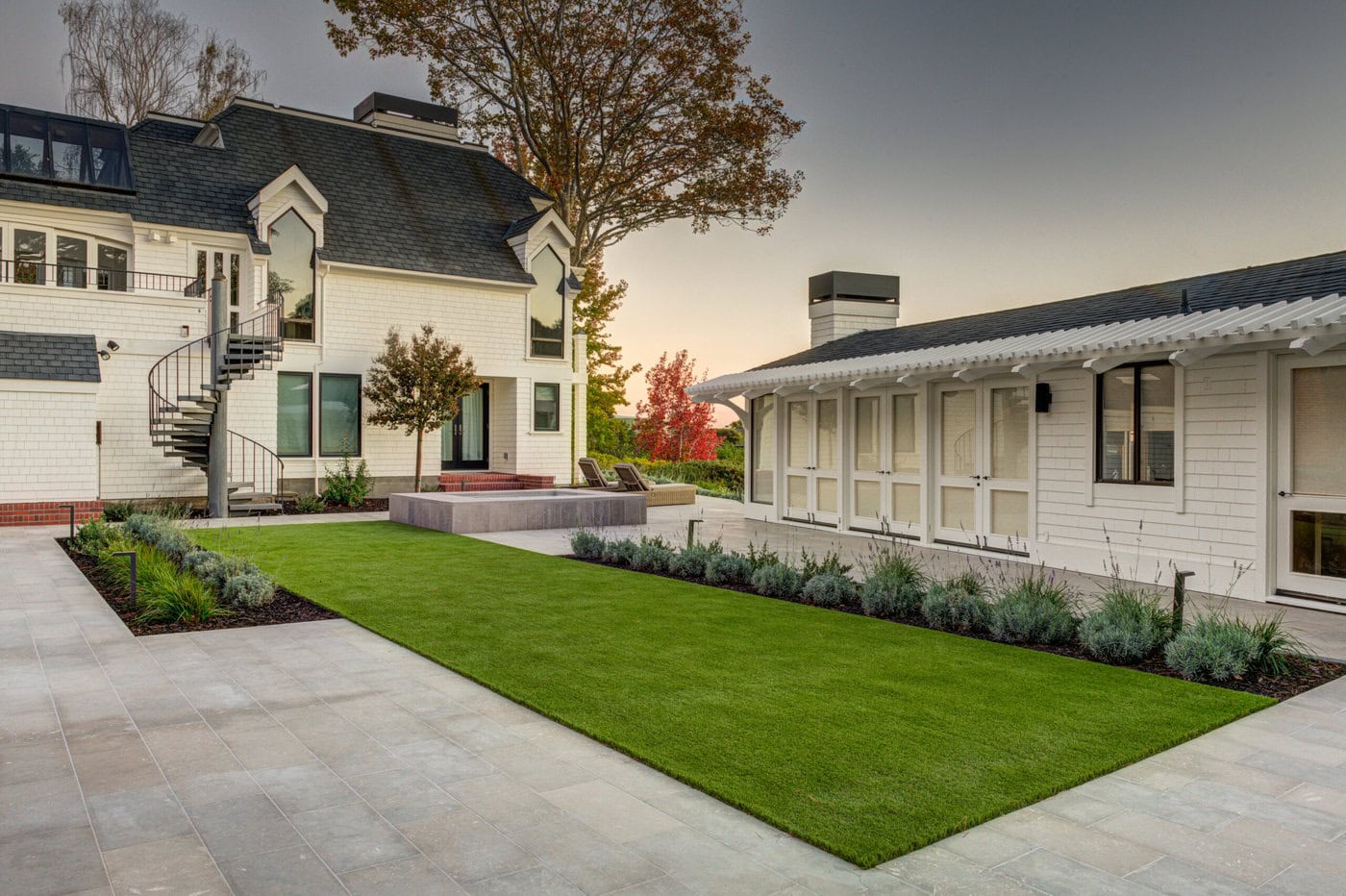 Modern House Exterior With White Siding, Dark Roof, And A Spiral Staircase Leading To The Upper Level. The Courtyard Features A Rectangular Lawn Bordered By Plants, And A Paved Area. A Sunset Sky Is Visible In The Background, Enhancing The Serene Setting.