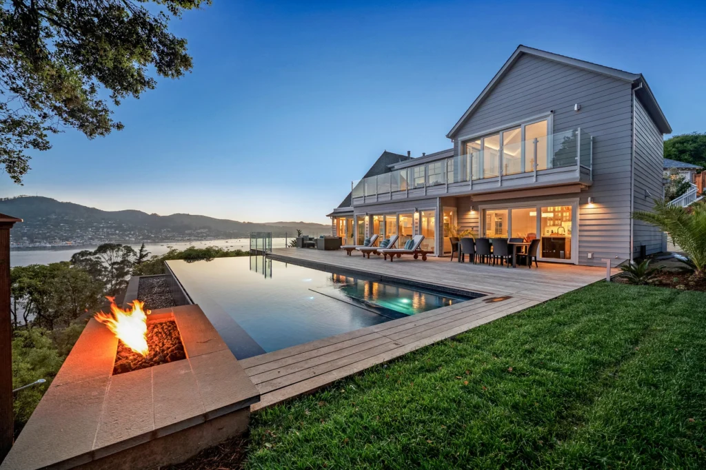 A Modern Two-Story House With Large Windows And A Deck Is Shown At Dusk. An Infinity Pool With Adjacent Lounge Chairs Overlooks A Scenic View Of Hills And Water. A Small Fire Pit Sits In The Foreground, Enhancing The Serene Atmosphere.